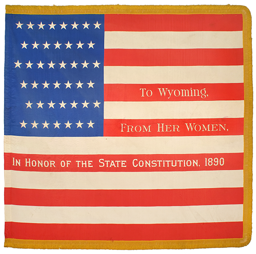 U.S. flag with To Wyoming from her women, in honor of the state constitution, 1890, written along the red stripes.  There are 44 stars